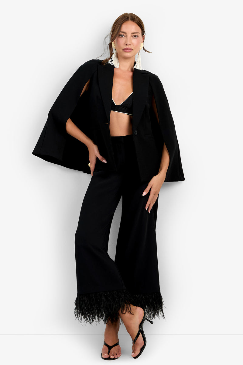 A woman wearing the Volterra Jacket—a black cape blazer—paired with a black bralette, wide-leg black pants with feathered hems, black heels, and white statement earrings stands against a plain white background.