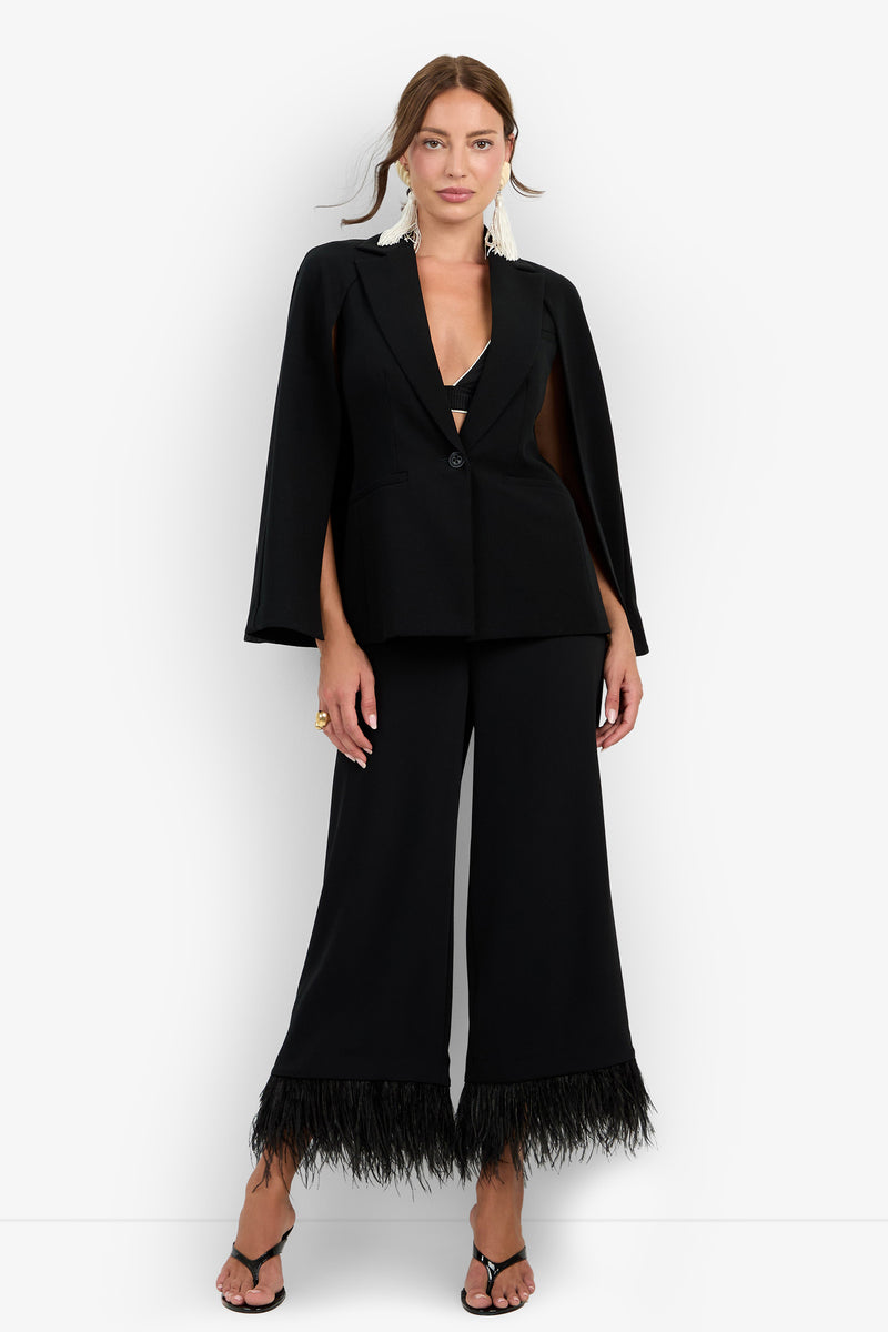 A woman stands against a plain white background wearing the Volterra Jacket in black—a cape-style blazer paired with wide-leg pants featuring feathered hems. She completes the look with black open-toe heels and large white statement earrings.