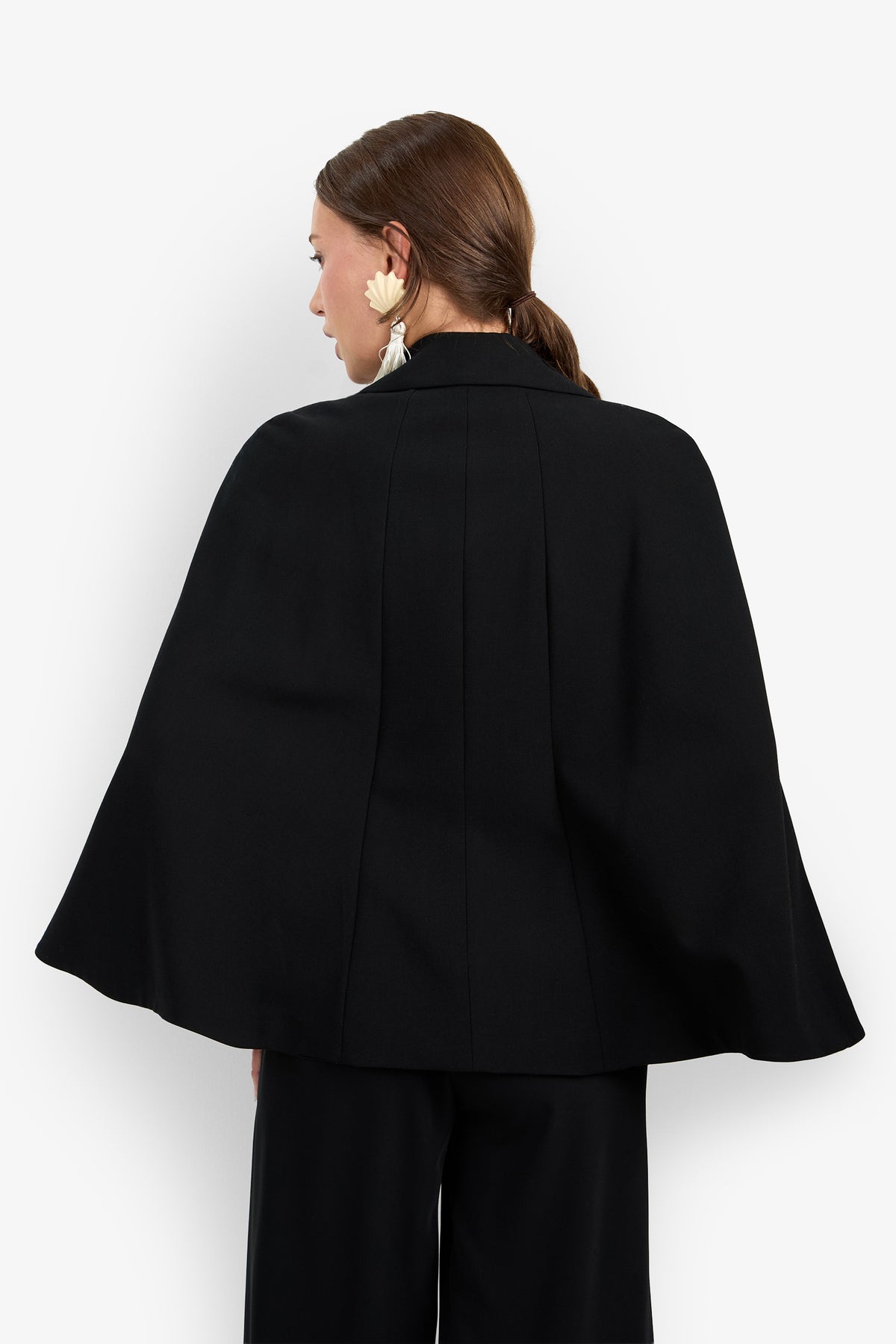 A woman with brown hair styled in a ponytail is seen from behind, wearing the Volterra Jacket—a black, cape-style piece—paired with wide-leg pants. She has on large floral earrings and is standing against a plain white background.