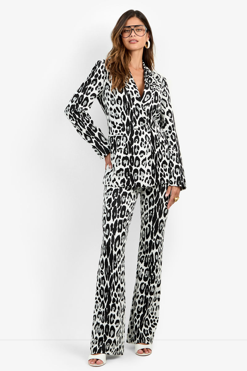 A woman wearing glasses and the Bacino Jacket in Snow Leopard, styled with matching flared pants and open-toed heels, stands confidently against a white background.