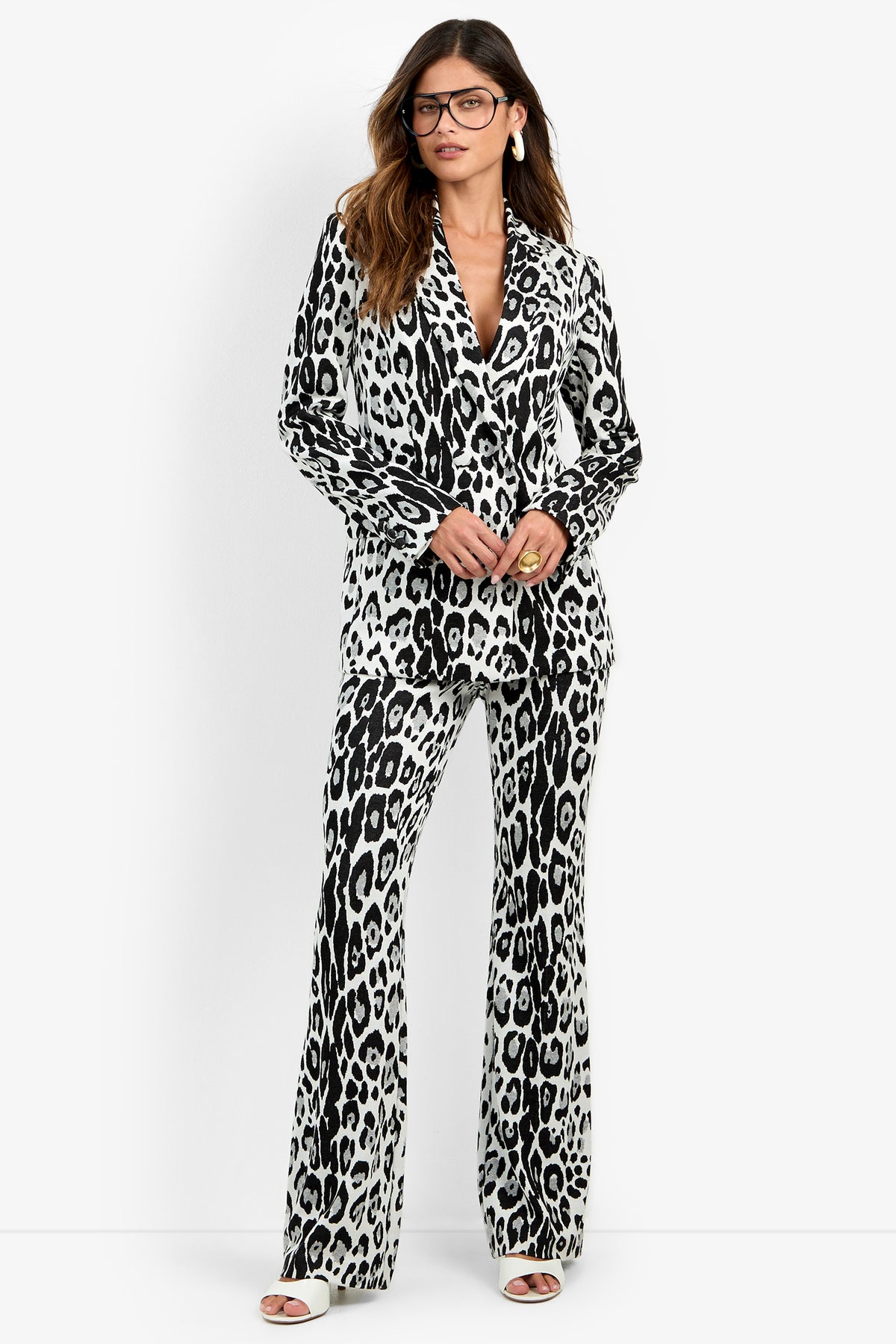 A woman wearing the Bacino Jacket, styled with black and white leopard print pants, white sandals, and glasses, stands against a white background with her hands clasped and looking forward.
