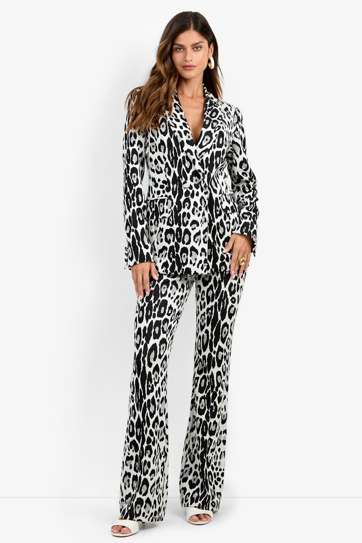 A woman stands wearing the Bacino Jacket—a black and white leopard-print double-breasted blazer styled with matching flared pants and white open-toed heels. She has wavy brown hair and accessorizes with hoop earrings.