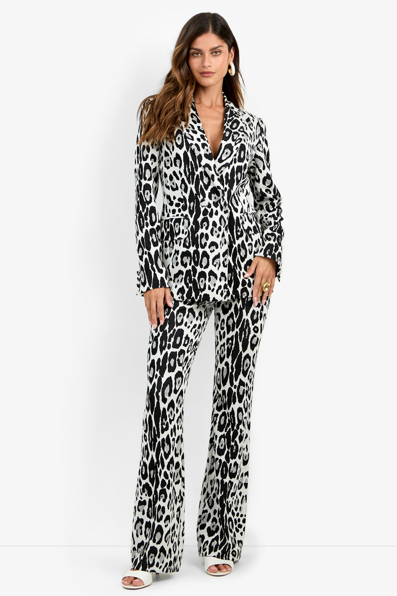 A woman stands wearing the Bacino Jacket—a black and white leopard-print double-breasted blazer styled with matching flared pants and white open-toed heels. She has wavy brown hair and accessorizes with hoop earrings.