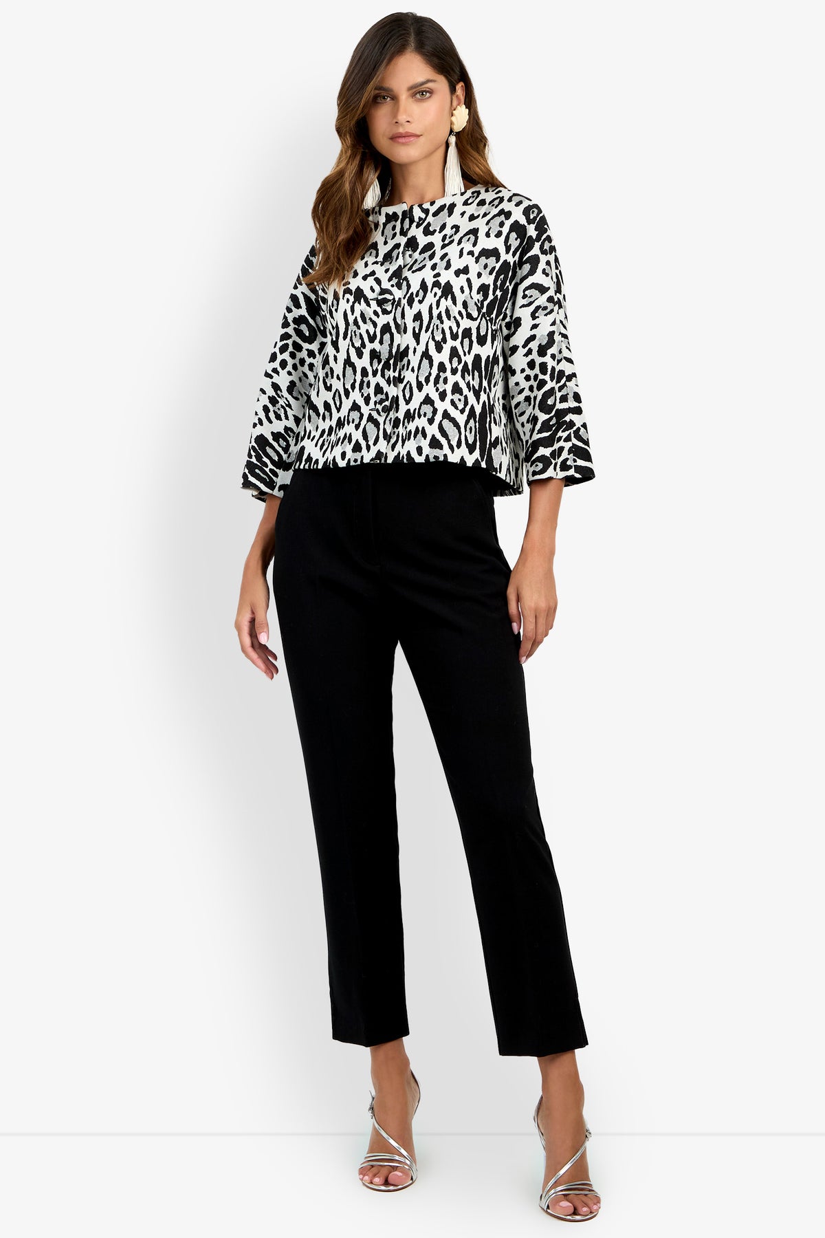 A woman wearing the Critique Jacket—a black and white leopard-print style—paired with black pants and silver high-heeled sandals, standing against a white background.