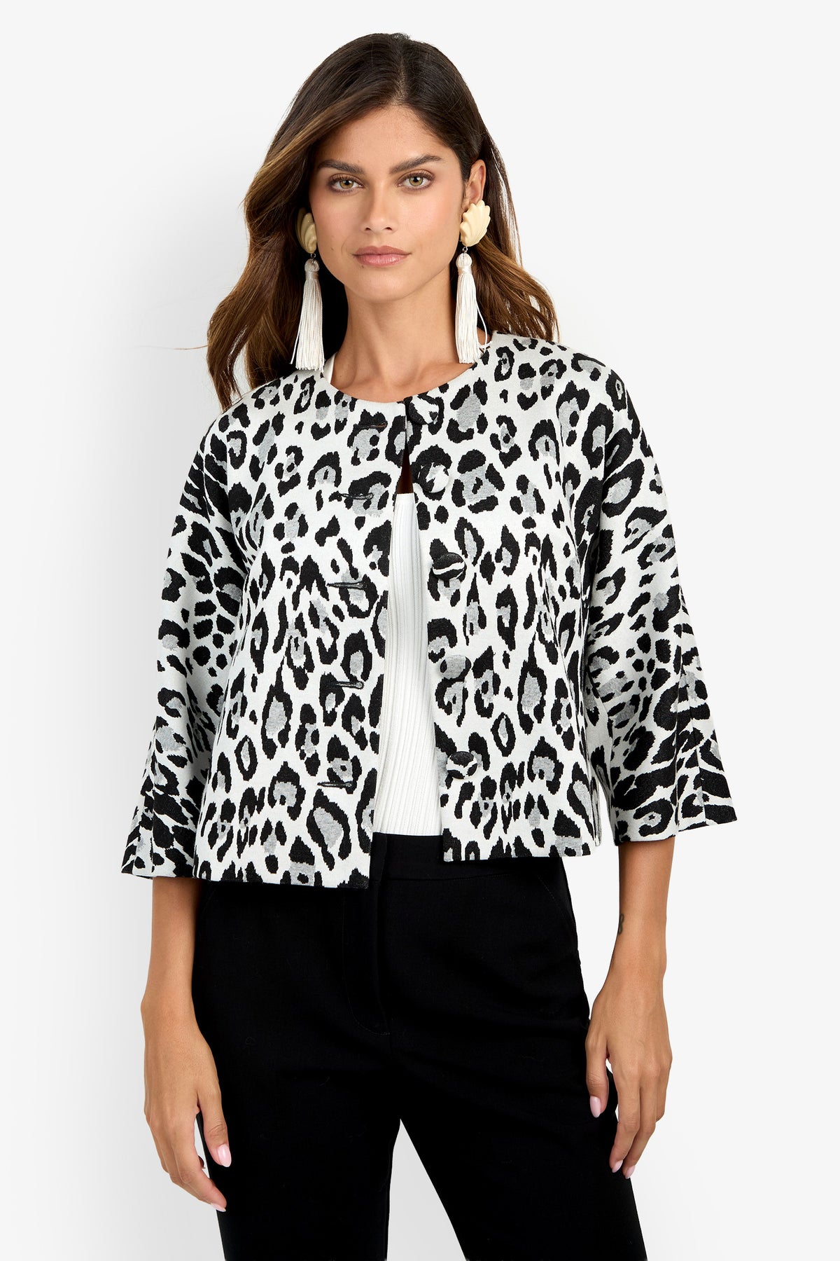 A woman stands against a plain white background, wearing the Critique Jacket—a black and white leopard print style—layered over a white top and black pants, accessorized with long tassel earrings.