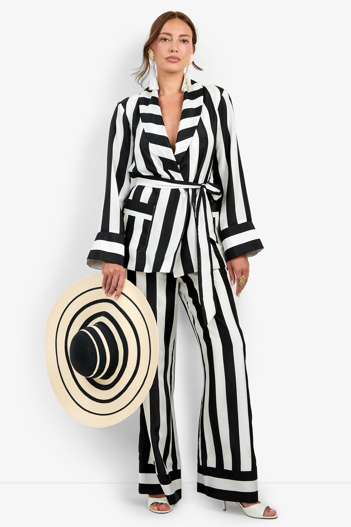 A woman wearing the Rolynn Jacket—a black-and-white vertically striped pantsuit—holds a large wide-brim hat featuring matching black concentric circle patterns, while standing against a plain white background.