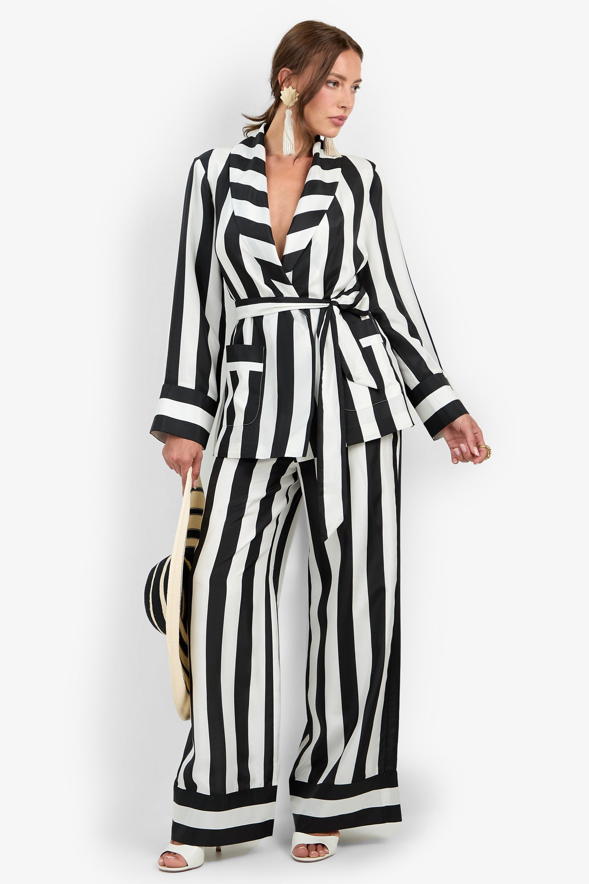 A woman wearing the Rolynn Jacket—a black and white vertically striped, belted blazer—styled with wide-legged matching pants, white heels, large earrings, and holding a striped hat.