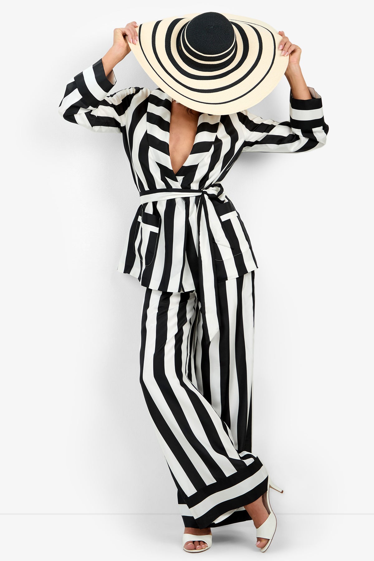 Wearing the Rolynn Jacket, a person poses with one hand holding the brim of their wide-brimmed black and white striped hat, face obscured. They stand against a plain white background in white heels.