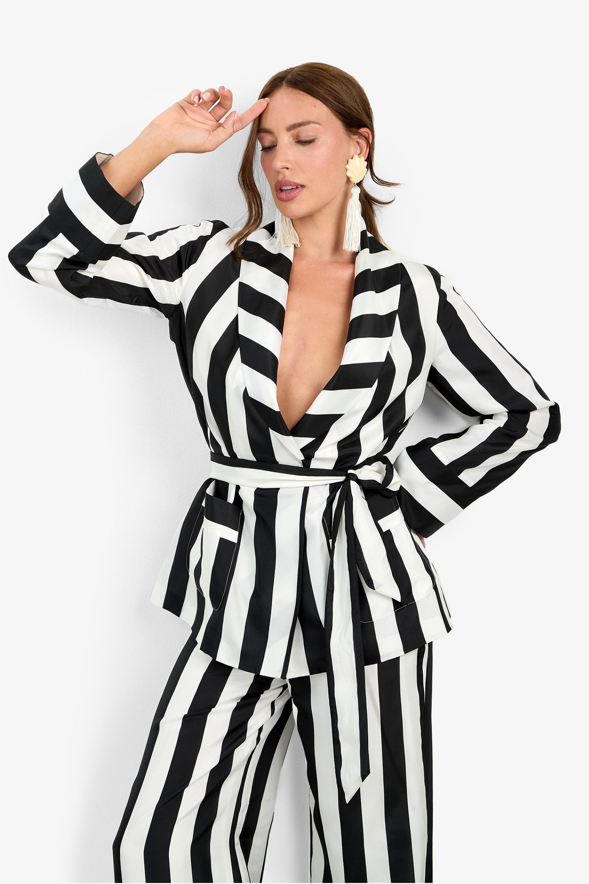 A woman models the Rolynn Jacket—a black-and-white vertically striped suit jacket with a belted waist—paired with wide-leg pants and large tasseled earrings, posing with one hand on her head and her eyes closed.