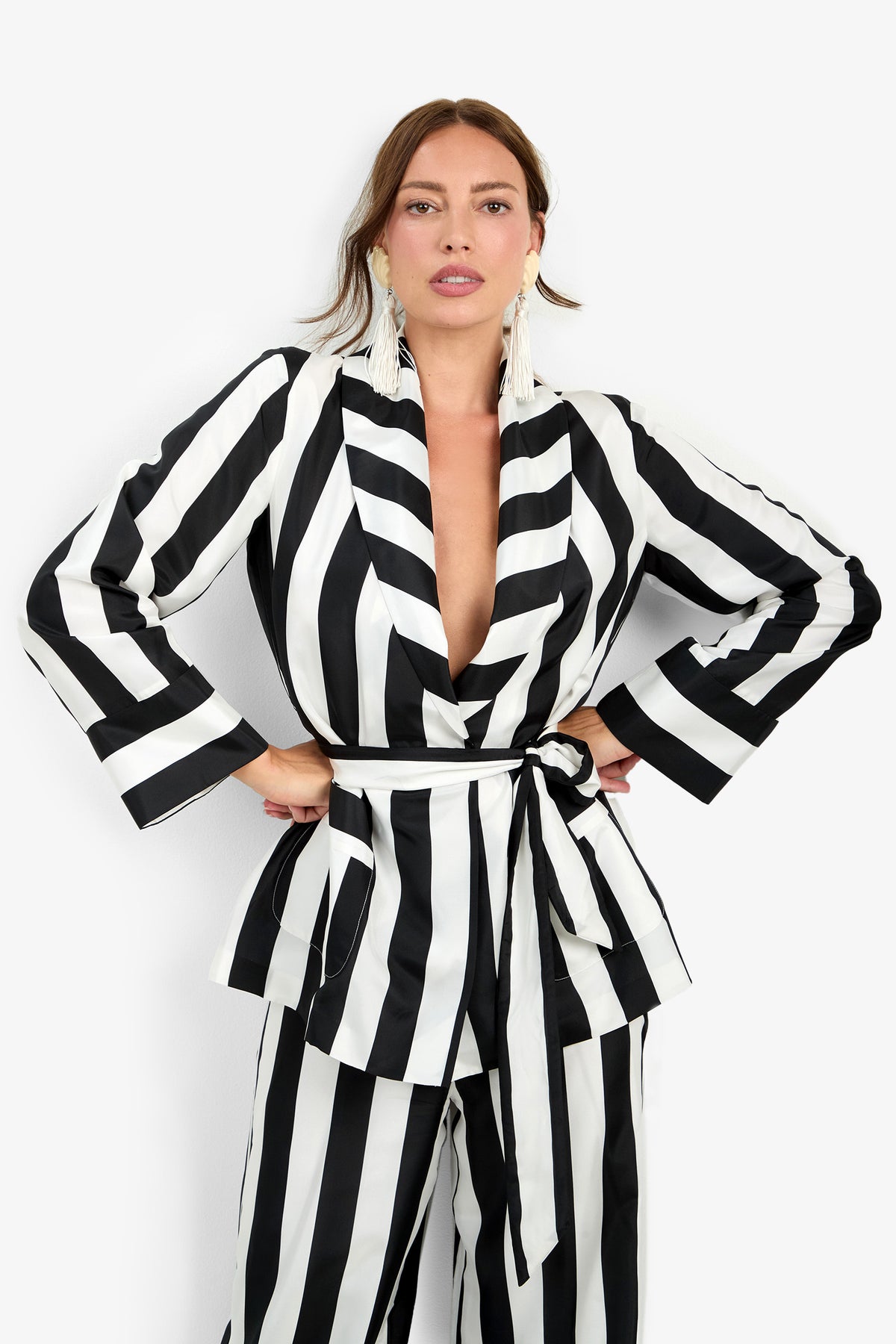 A woman wearing the Rolynn Jacket—a black and white vertical striped suit featuring a belted waist, wide sleeves, and matching pants. She accessorizes with tassel earrings and stands with hands on hips against a white background.