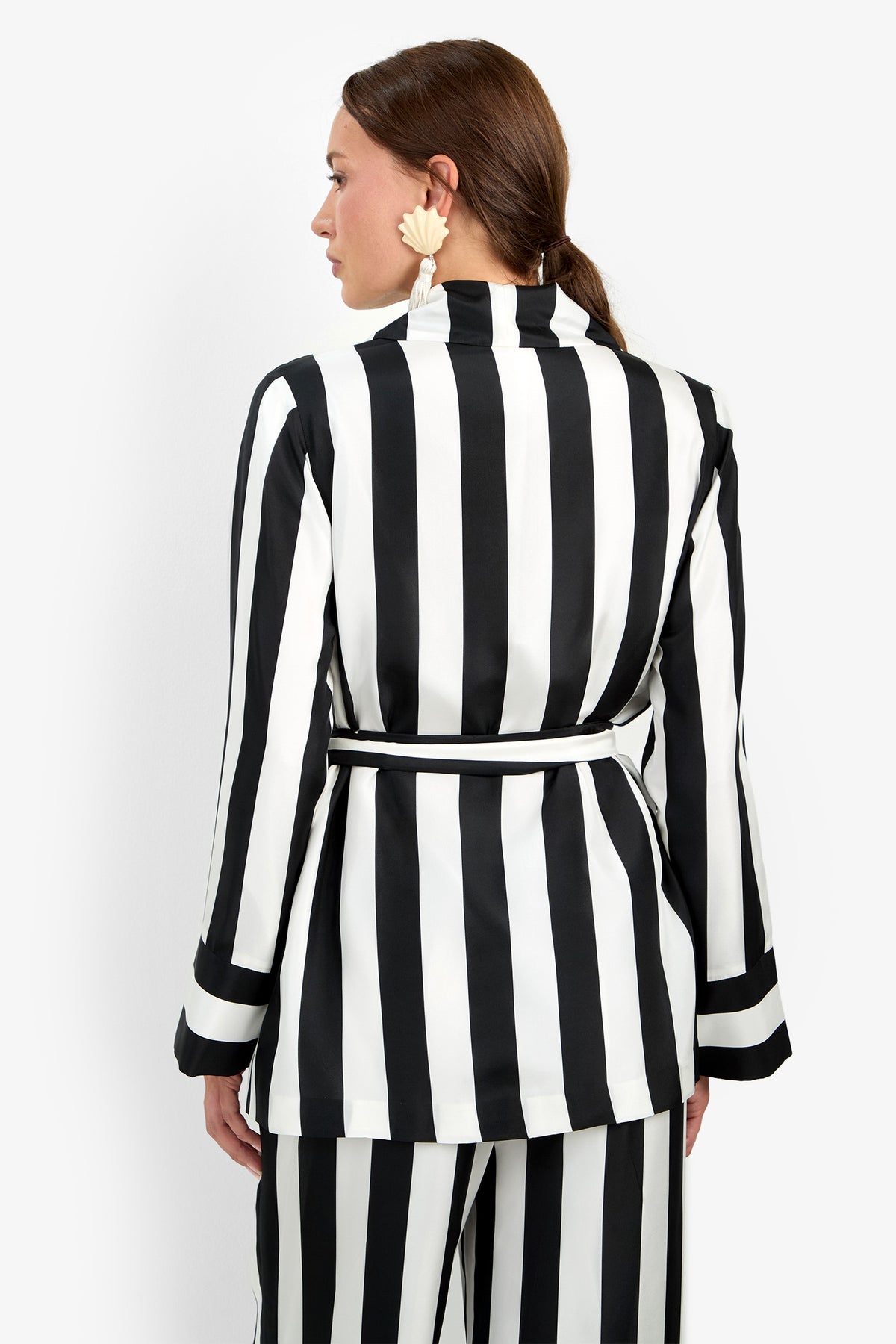 A person stands facing away, wearing the Rolynn Jacket—a black and white vertically striped suit with a belted waist. Their hair is tied back, and they accessorize with large, light-colored floral earrings.