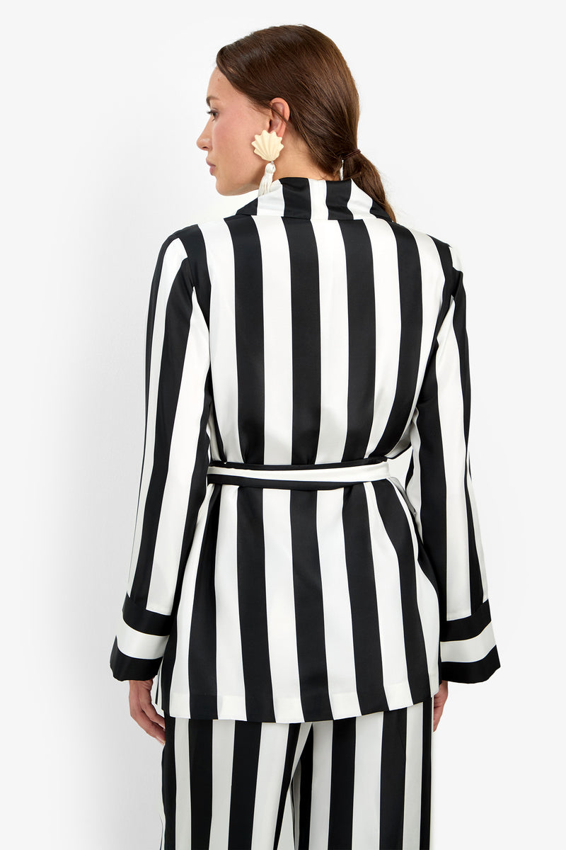 A person stands facing away, wearing the Rolynn Jacket—a black and white vertically striped suit with a belted waist. Their hair is tied back, and they accessorize with large, light-colored floral earrings.
