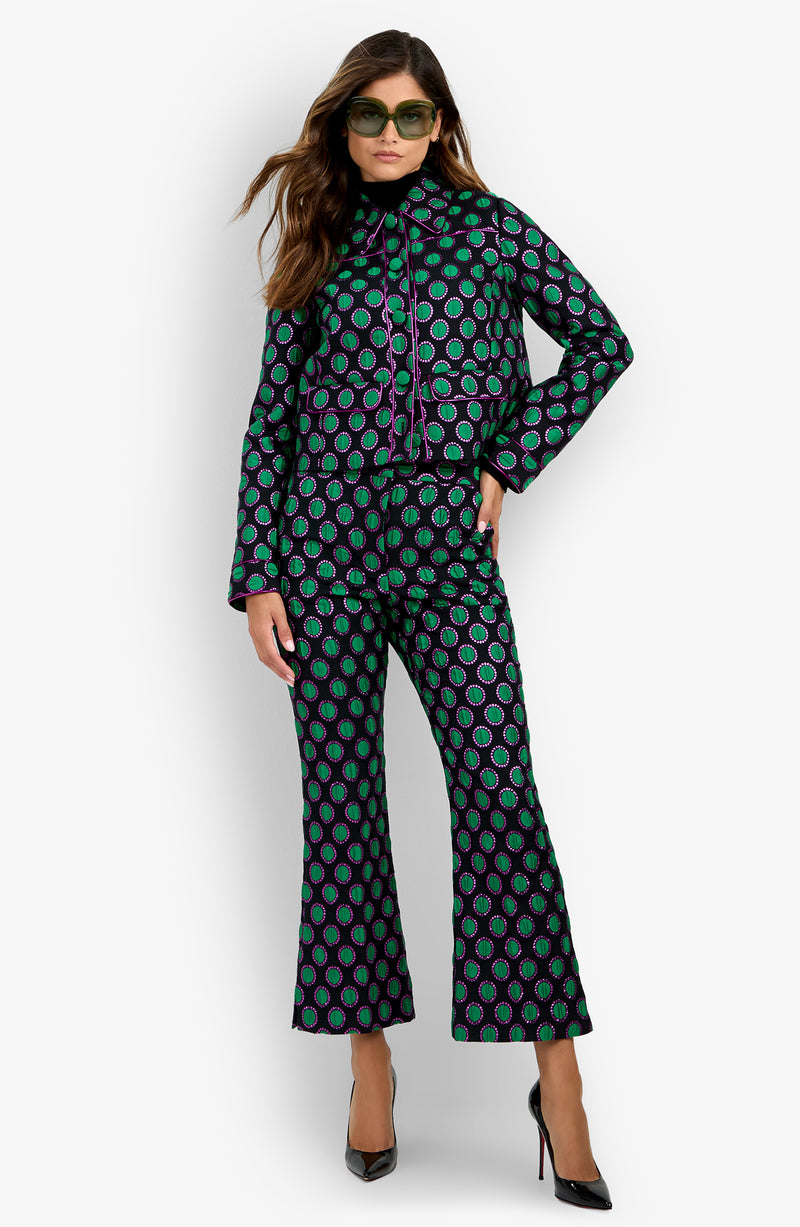 Woman wearing the Allex Jacket styled with matching flared pants featuring a geometric green and purple pattern, black high heels, and sunglasses, standing against a white background.