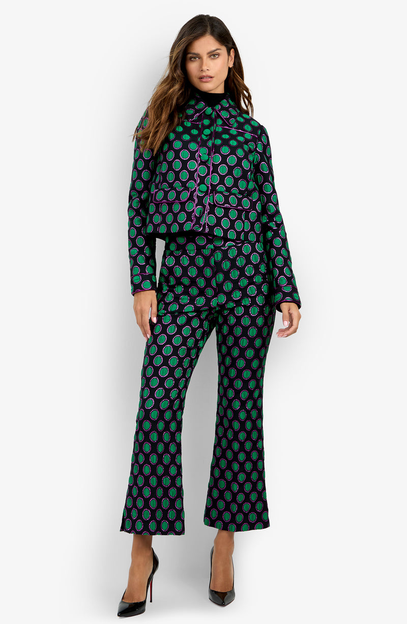 A woman standing against a white background wears the Allex Jacket—a cropped black jacket with green and purple circular patterns—paired with matching flared pants and black heels.
