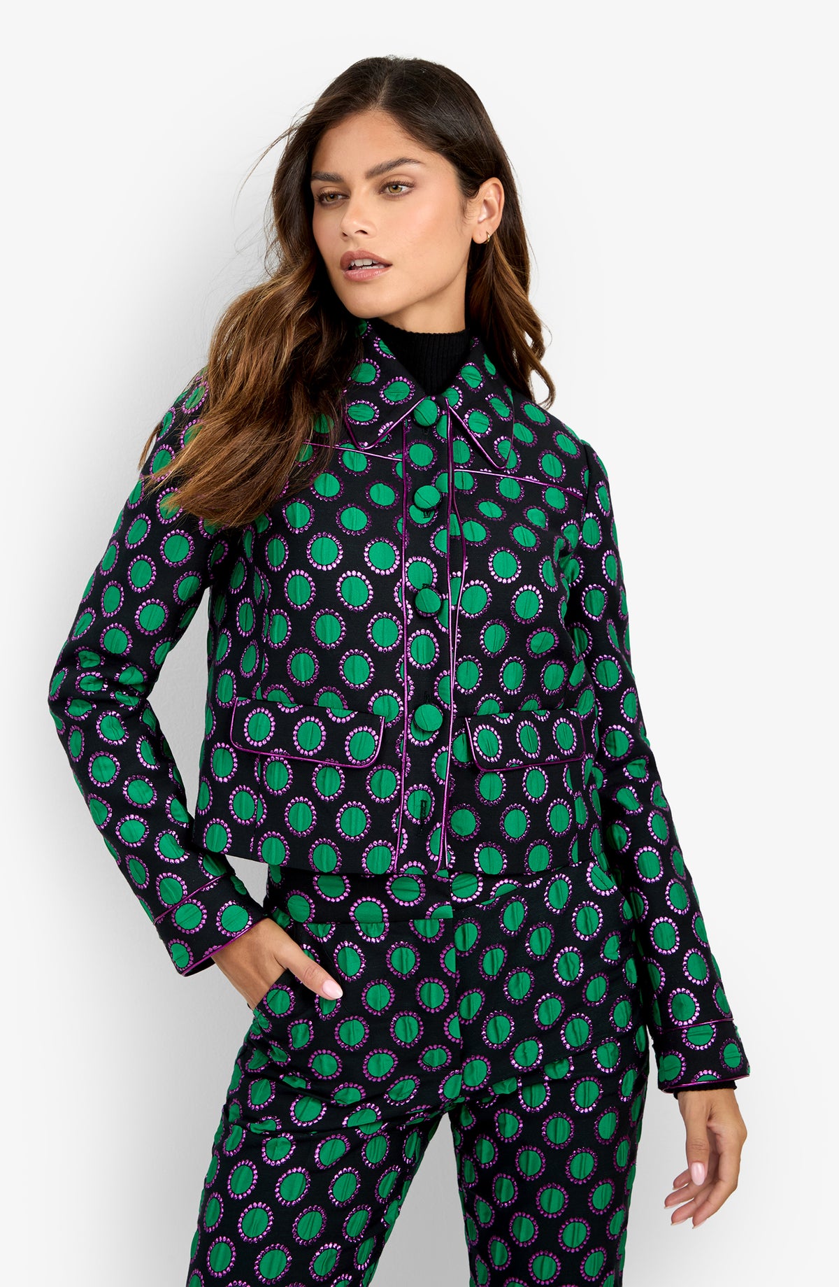 A woman poses with one hand in her pocket against a white backdrop, wearing the Allex Jacket—a matching set featuring a green and purple circular pattern on a black background.
