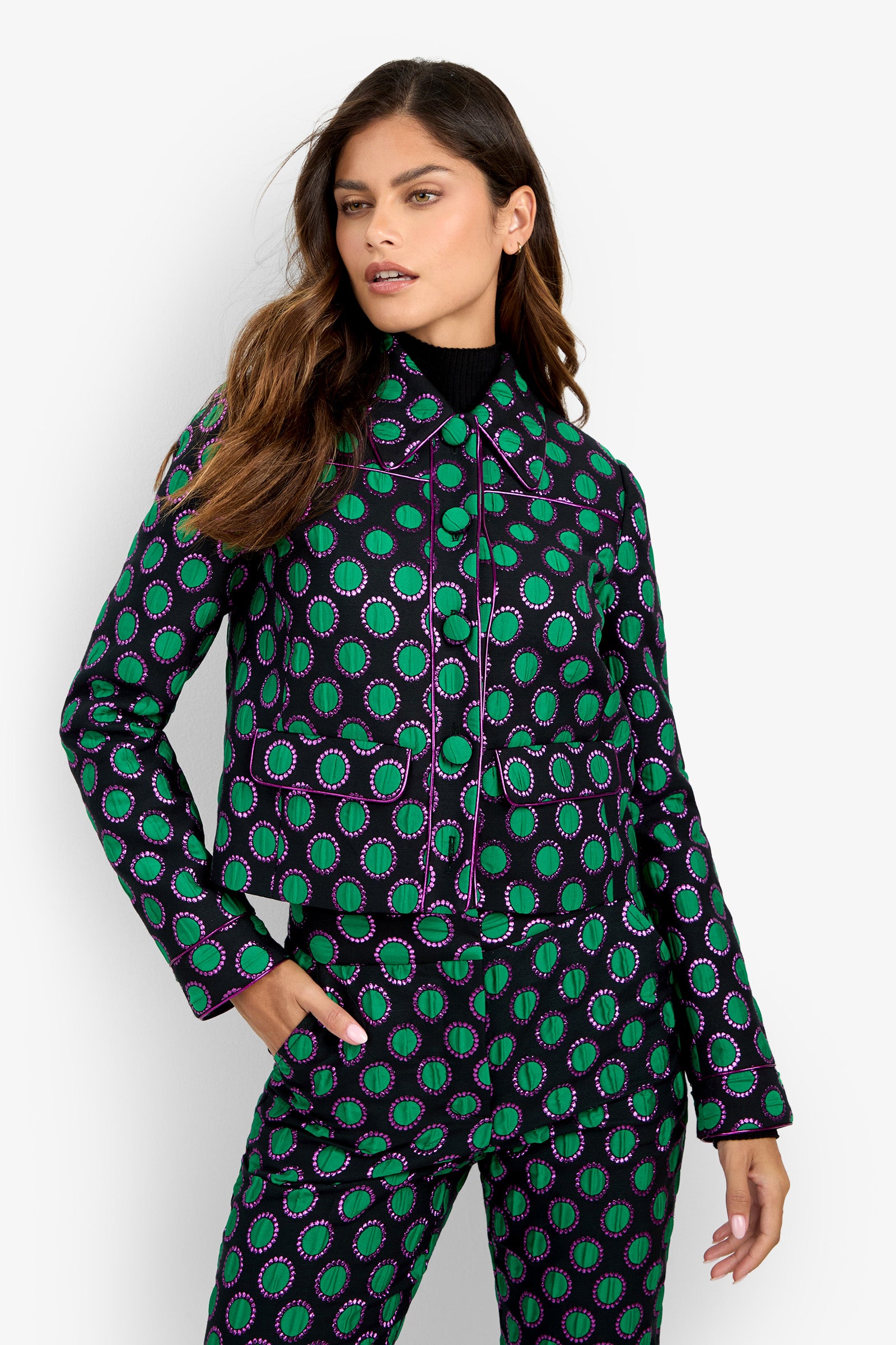 A woman poses against a plain white background wearing the Allex Jacket, featuring black, green, and purple circular patterns, paired with matching pants and a black turtleneck underneath.