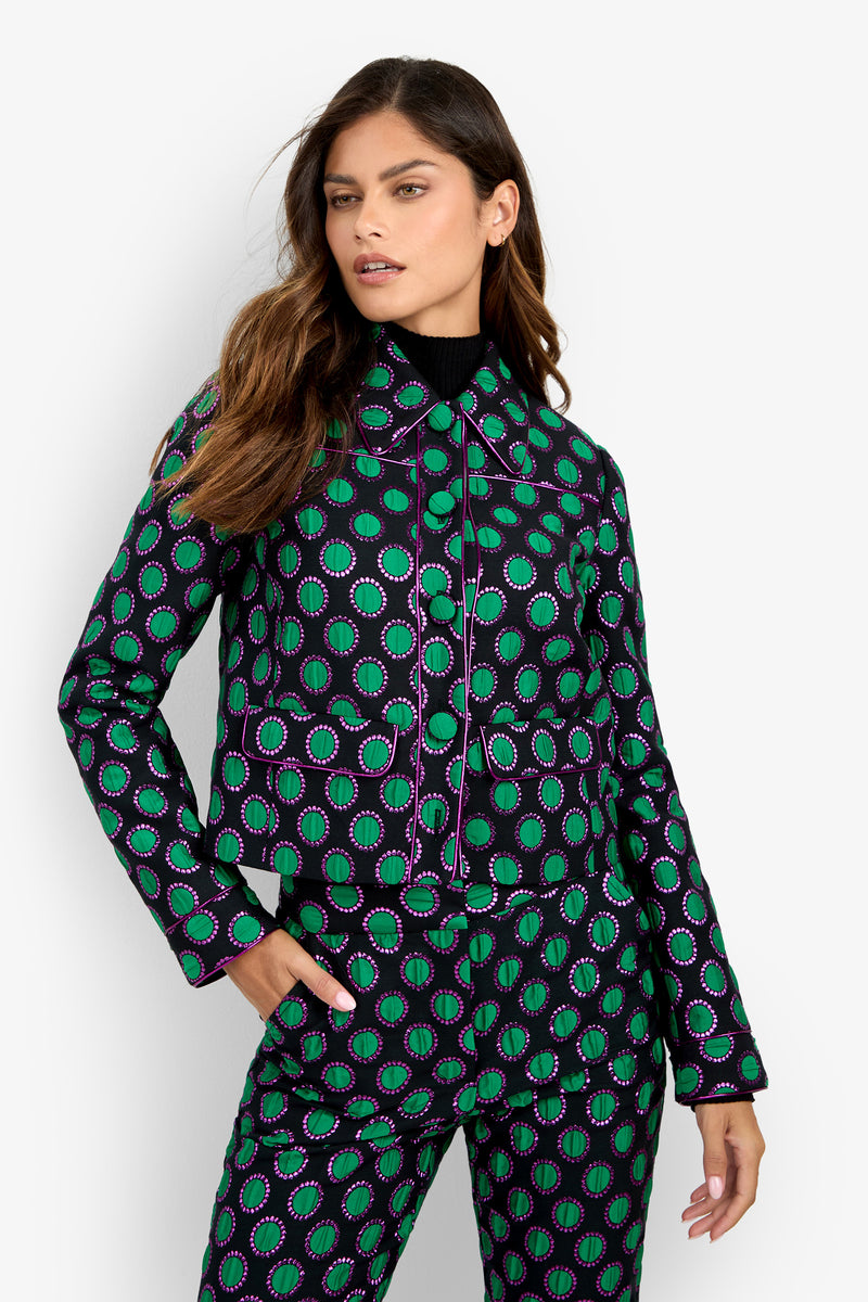A woman poses against a plain white background wearing the Allex Jacket, featuring black, green, and purple circular patterns, paired with matching pants and a black turtleneck underneath.