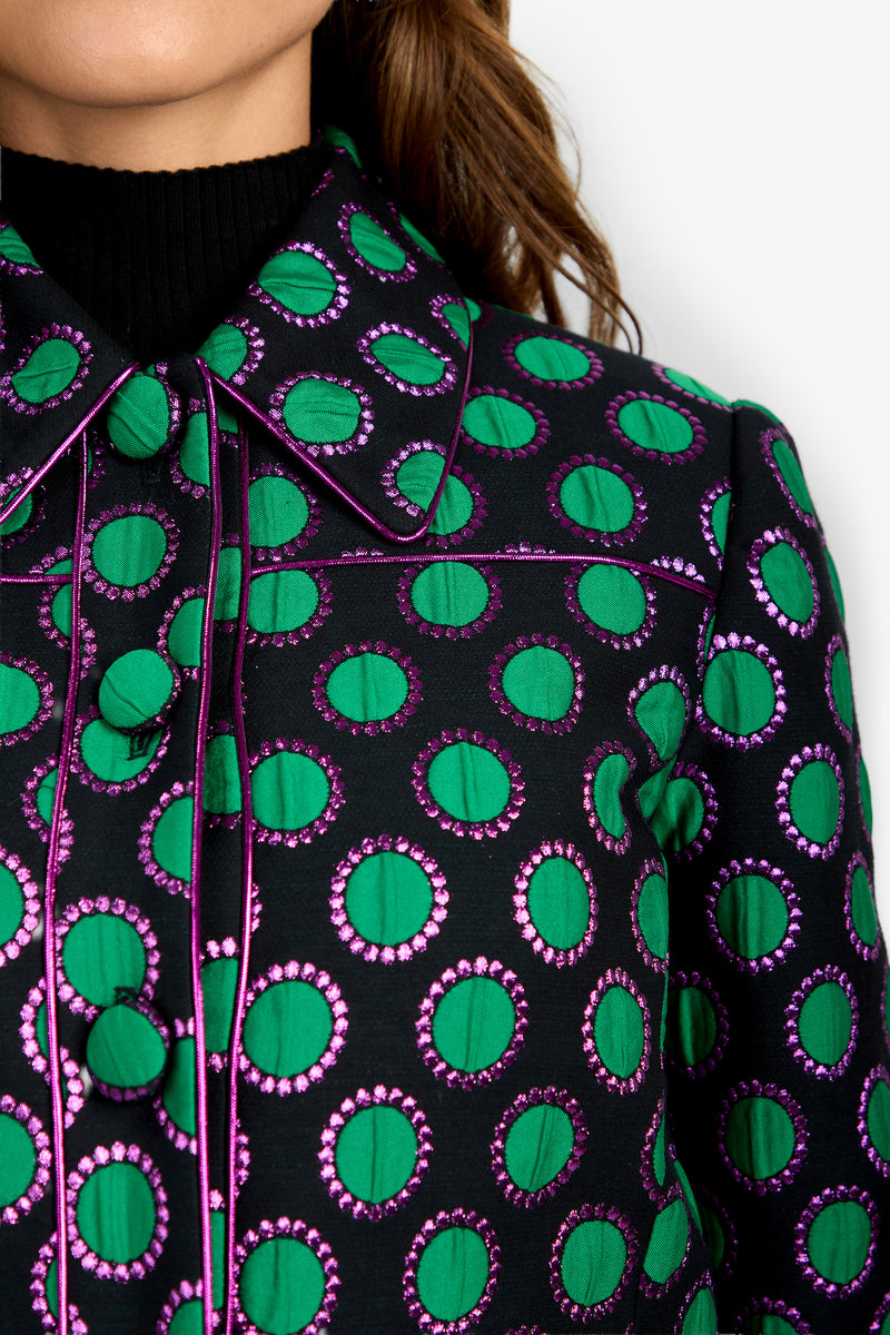 A close-up of a woman’s torso wearing the Allex Jacket, a black shirt featuring green and purple circular patterns with a pointed collar, layered over a black ribbed turtleneck.