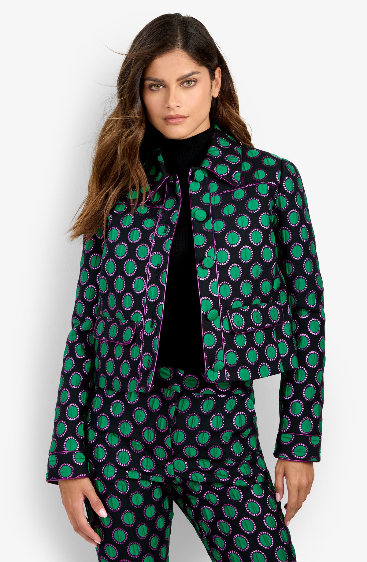 Woman in the Allex Jacket and matching pants, featuring green, purple, and white circular patterns, stands against a white background. She has long brown hair and wears a black turtleneck underneath.
