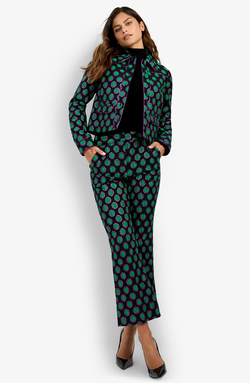 A woman stands confidently with her hands in her pockets, wearing the Allex Jacket—a matching patterned suit featuring green and purple circles—paired with a black top and black heels.
