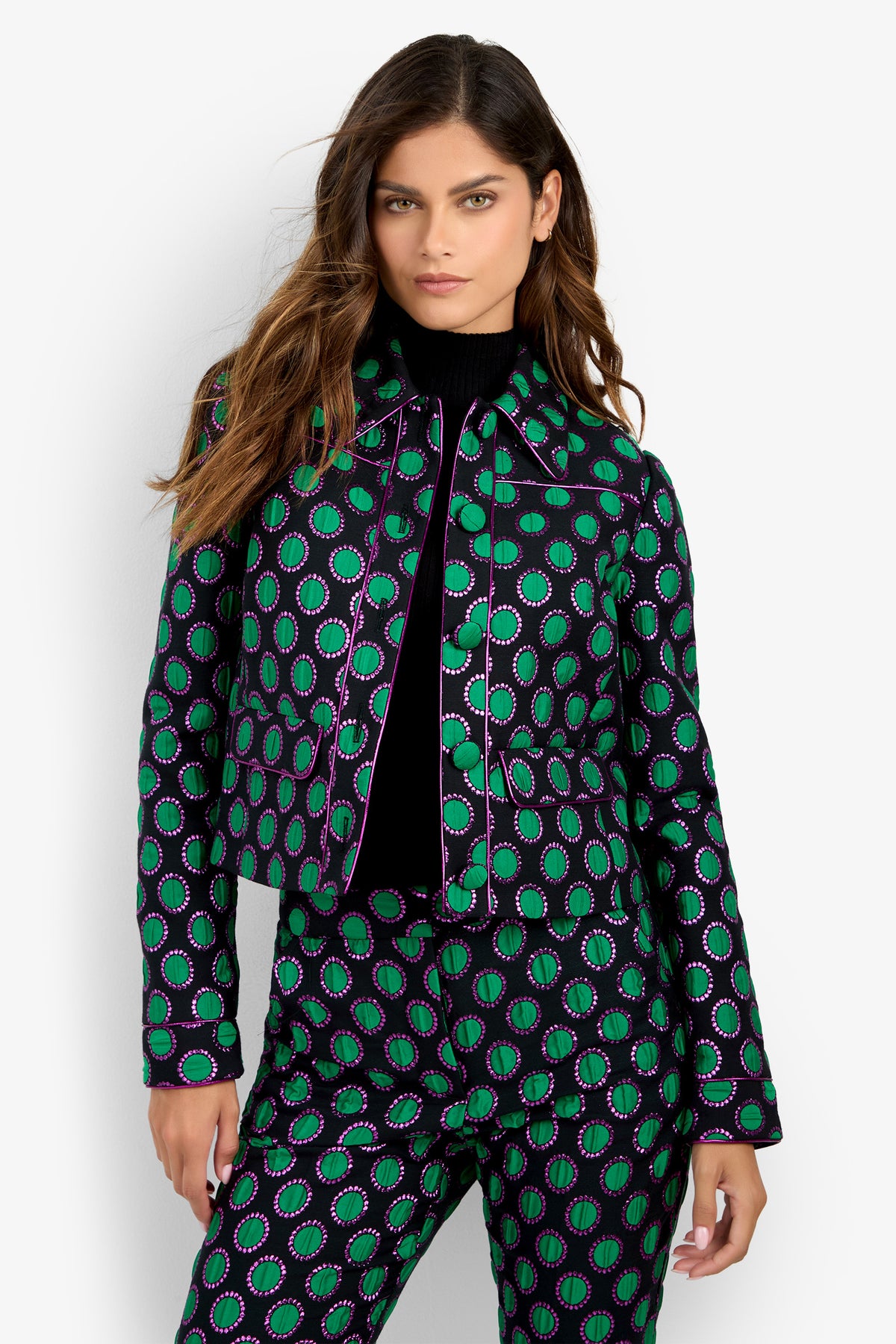 A woman with long brown hair wears the Allex Jacket—a black, green, and purple patterned design—paired with matching pants over a black turtleneck, standing against a plain white background.