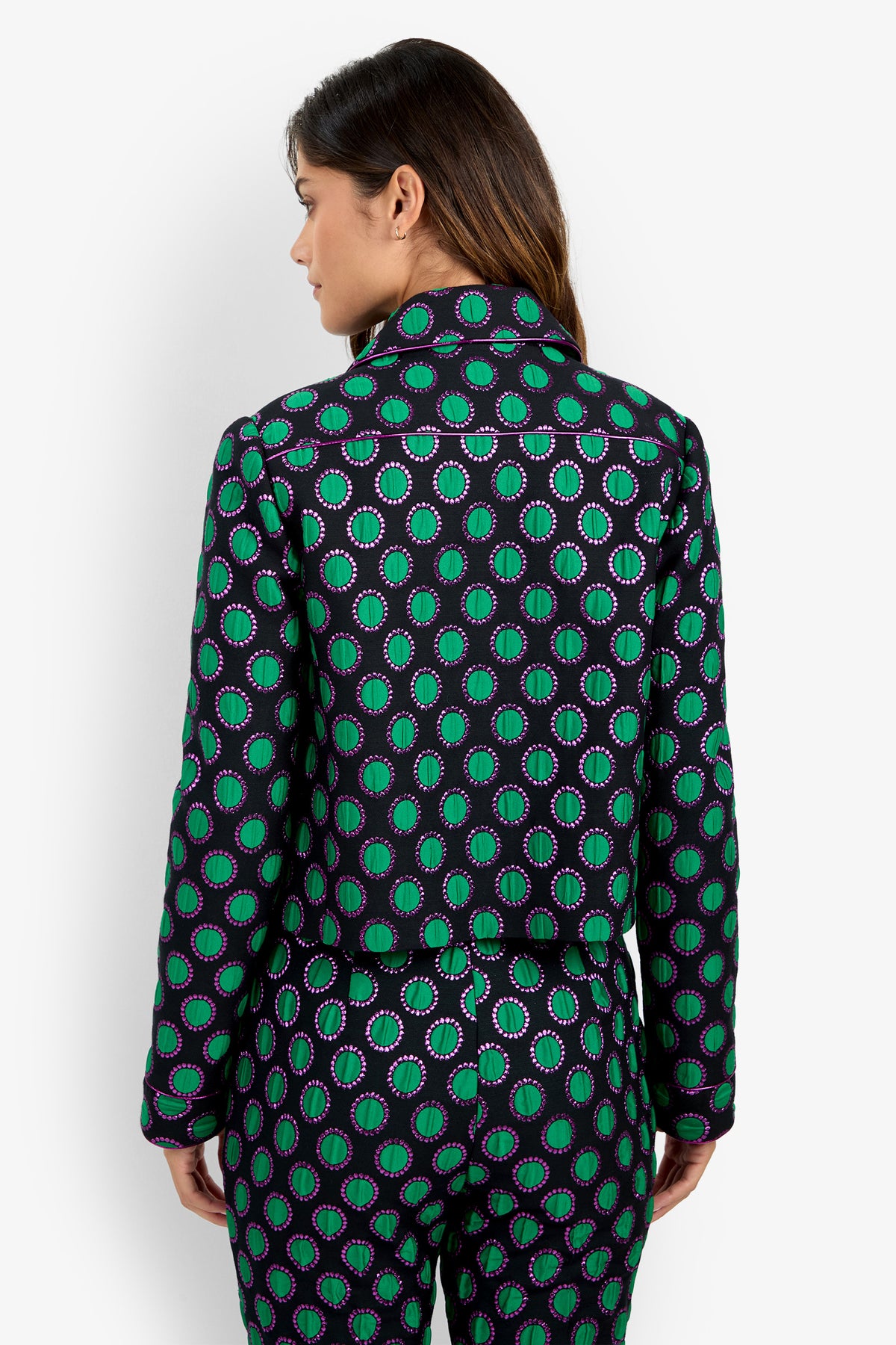 A woman faces away, wearing the Allex Jacket and matching pants featuring a vibrant green and purple circular pattern. Her brown hair is down, with a plain white background behind her.