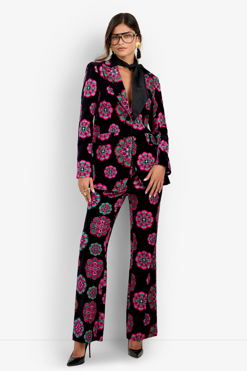 Wearing the Broadway Jacket—a velvet suit jacket adorned with colorful floral patterns—paired with a black necktie blouse, black heels, large glasses, and statement rings, a woman stands confidently against a white background.