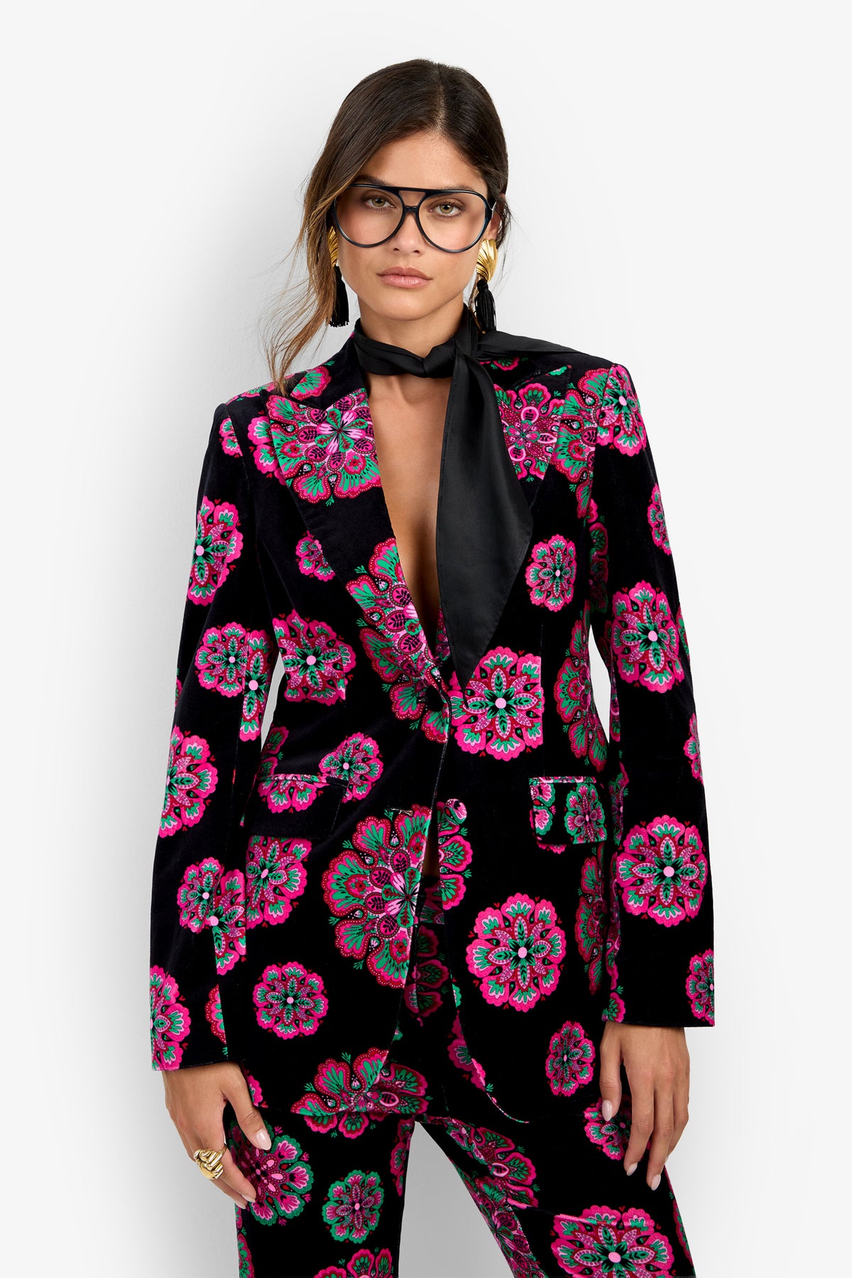 A woman wearing the Broadway Jacket—a black suit adorned with vibrant pink and green floral patterns—paired with gold hoop earrings, large black glasses, a black scarf, and multiple rings, stands against a plain white background.