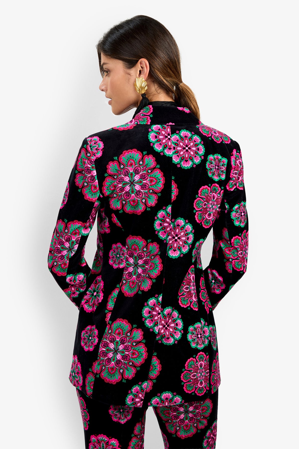 A woman with brown hair in a low ponytail wears the Broadway Jacket, a black blazer featuring bold pink, green, and white floral patterns. She faces away, showcasing the back of the blazer and its matching trousers.