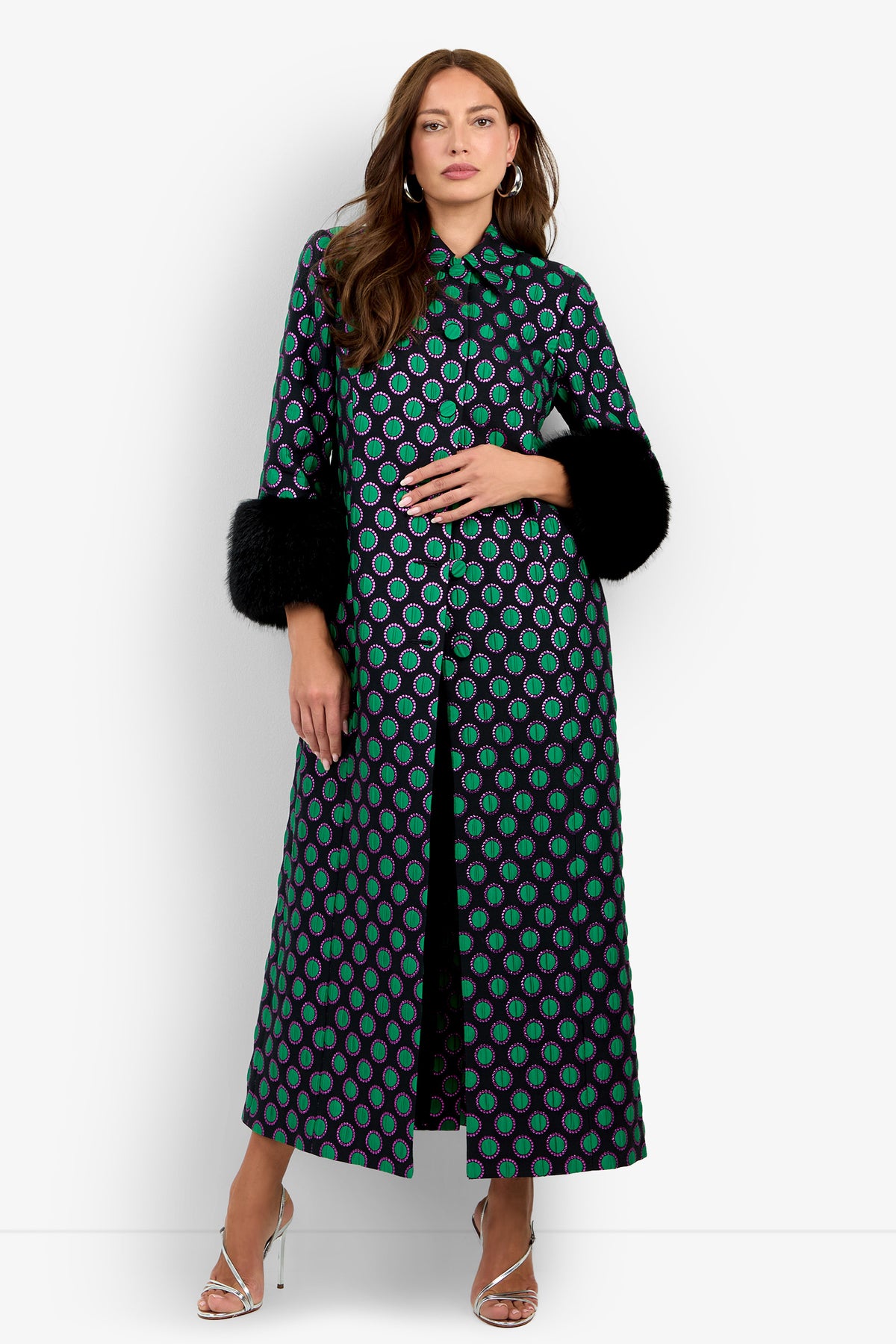 A woman wearing the Dalaine Coat—a long, patterned coat featuring green and purple circles with black fur cuffs—pairs it with silver heels and stands against a white background.