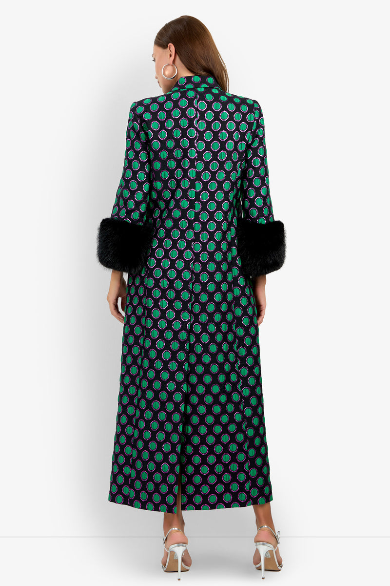 A woman stands with her back to the camera, wearing the Dalaine Coat—a long, patterned coat featuring green circles on a black background—styled with black faux fur cuffs, large hoop earrings, and white heeled sandals. The background is plain white.