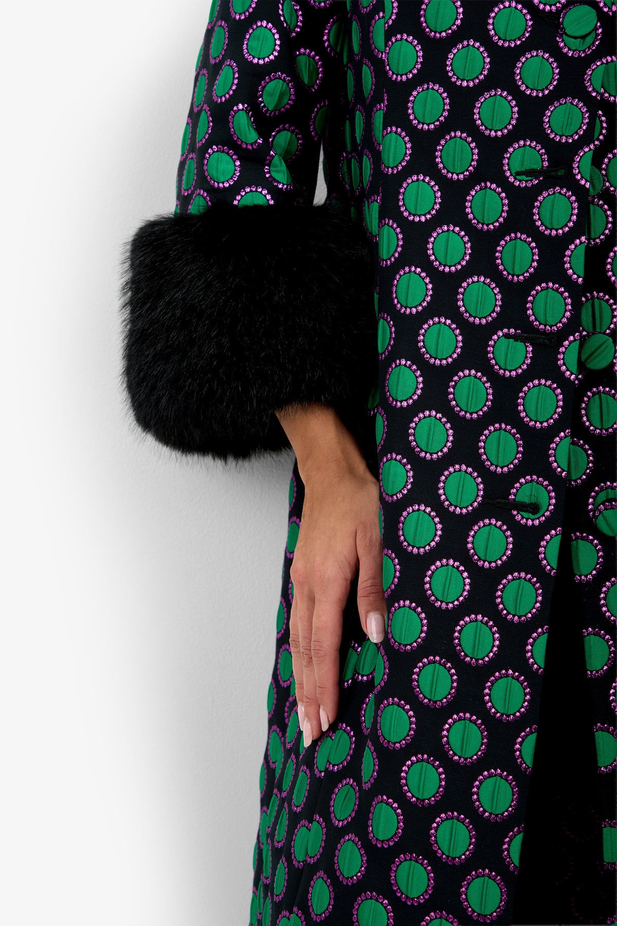 A person wearing the Dalaine Coat, featuring a green and purple pattern and black fur cuffs, with only their lower arm and hand visible against a white background.