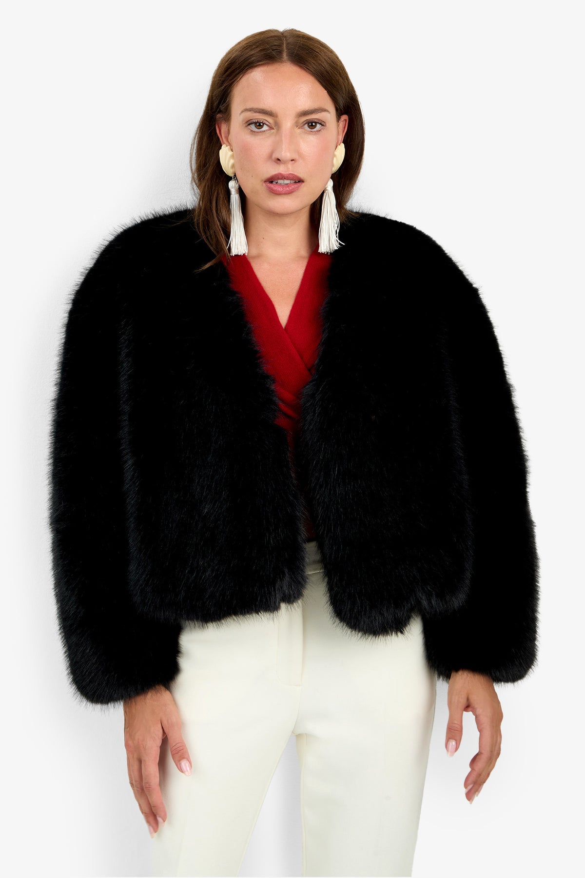 A woman wearing the Avenn Jacket, a black faux fur style, along with a red top, white pants, and white tasseled earrings stands facing forward against a plain background.