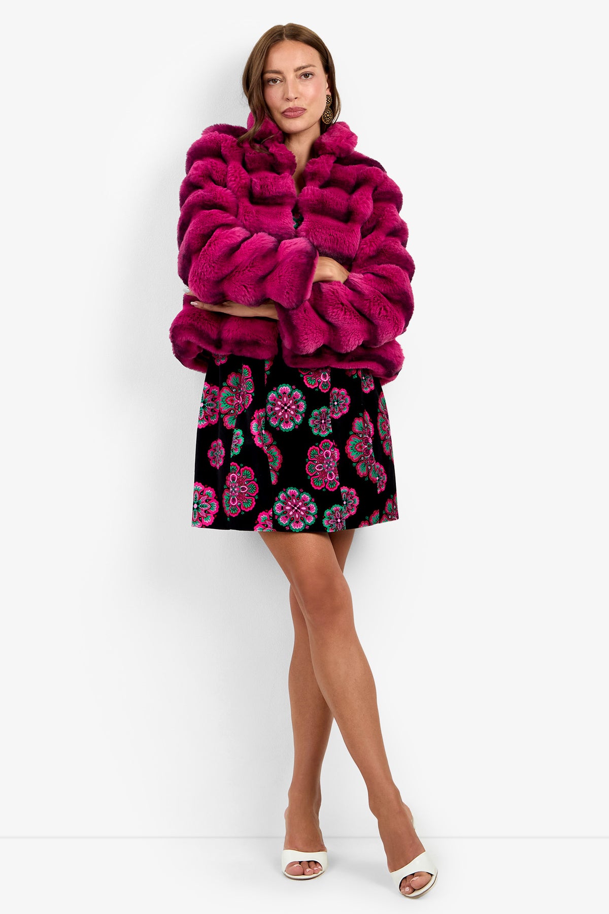 A woman wearing the Mikel Coat—a bright pink faux fur jacket—paired with a black skirt featuring colorful floral patterns and white heels stands against a white background with her arms crossed.