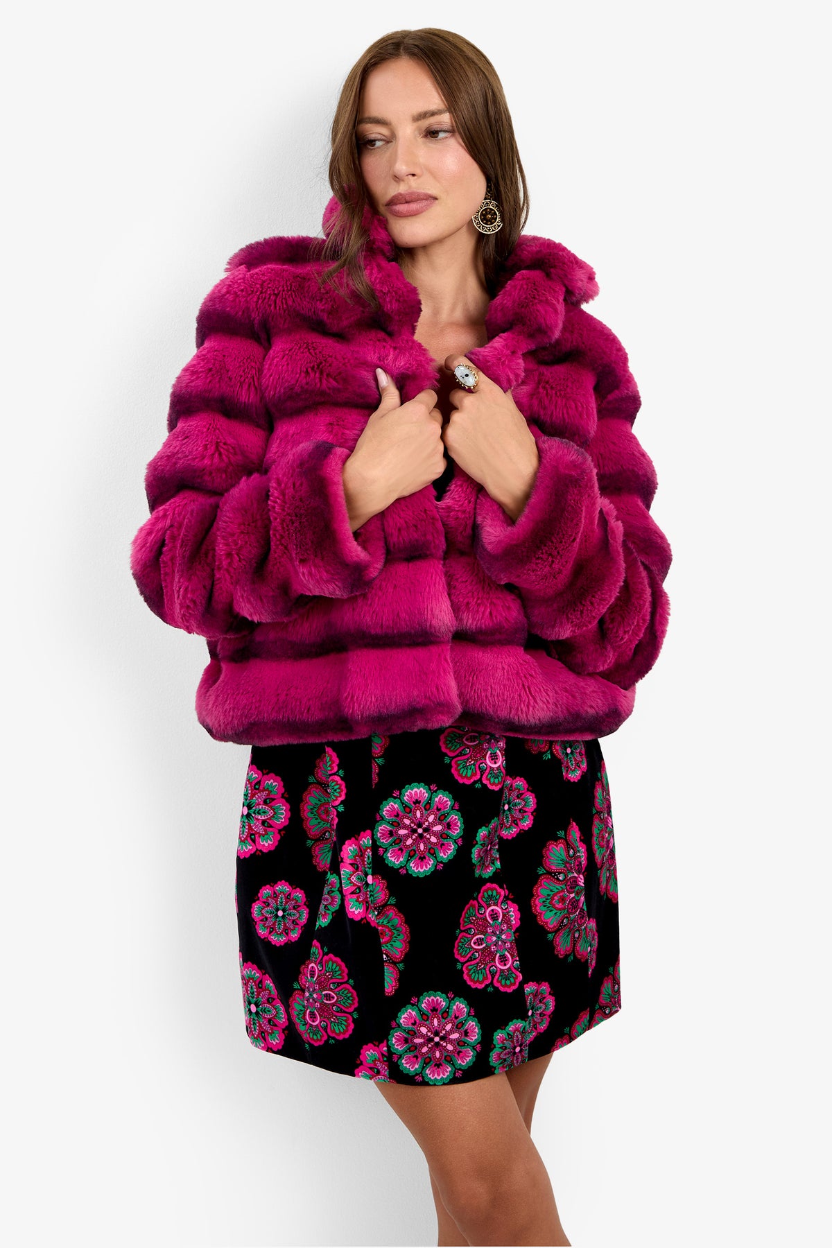 A woman stands facing forward, her hands holding the collar of the vibrant Mikel Coat—an eye-catching bright pink faux fur piece—paired with a black skirt adorned with pink and green floral patterns.