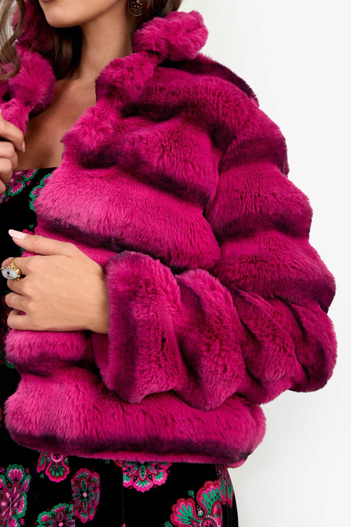 A woman wearing the Mikel Coat, a bright pink faux fur jacket, over a black dress with colorful floral patterns, holding the jacket collar with one hand.