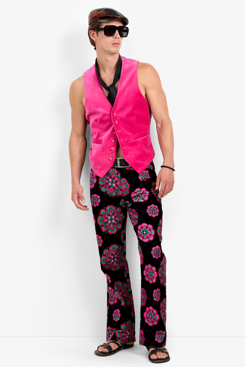 A man wearing a brown hat, sunglasses, the IRVING VEST in TORMALINA, a black scarf, floral-patterned spandex bell-bottom pants, a belt, and black sandals stands against a plain white background.