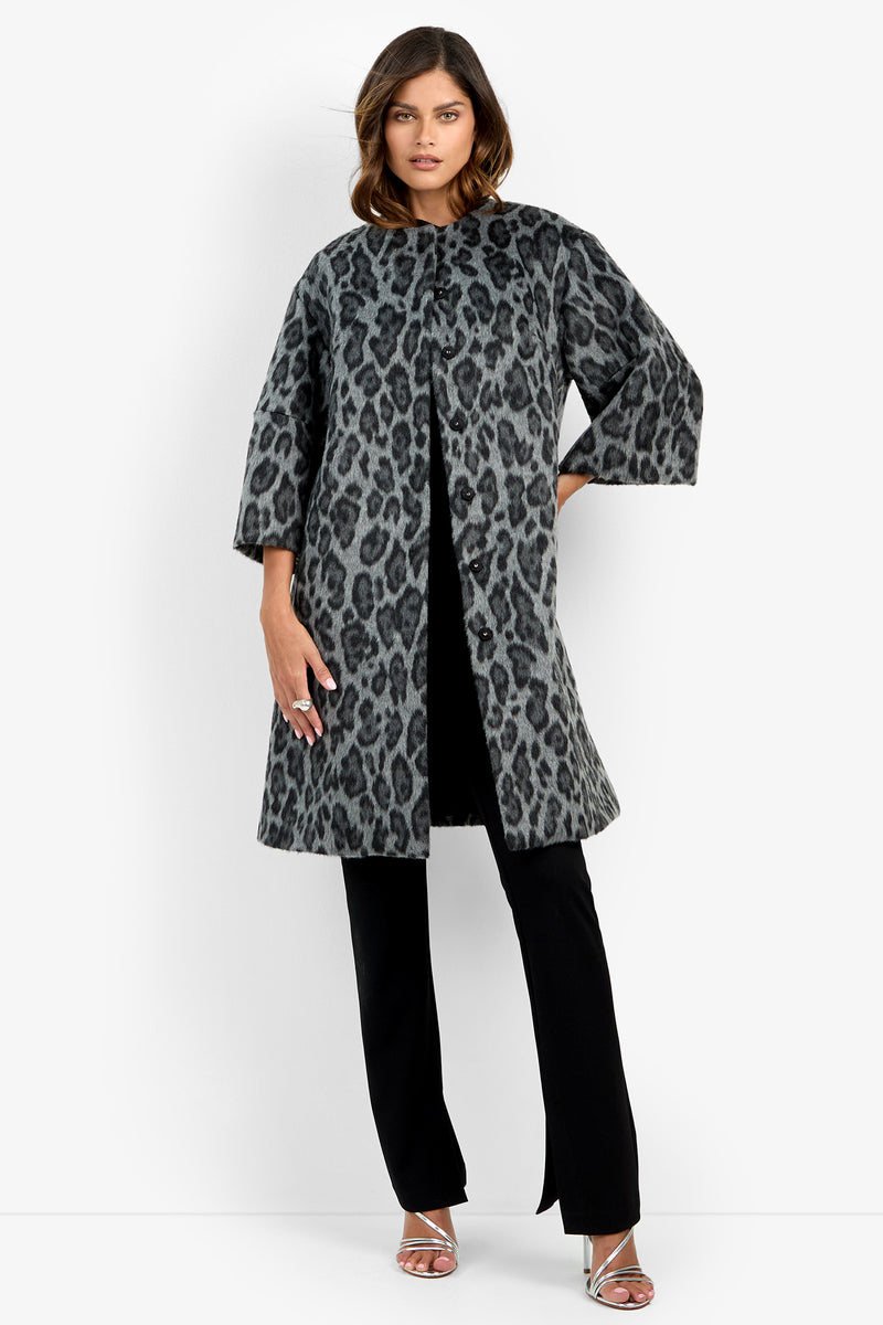 A woman wears the Lovely Leopard Coat in Snow Leopard—a knee-length coat with wide sleeves and a straight silhouette—over black pants and silver strappy heels, standing against a plain white background.