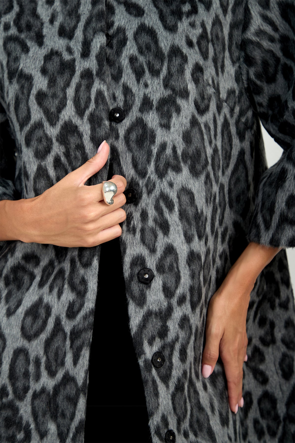Modeling the Lovely Leopard Coat—a chic gray and black leopard print design with black buttons—a person displays a striking large silver ring on their finger; only their hands and torso are visible.