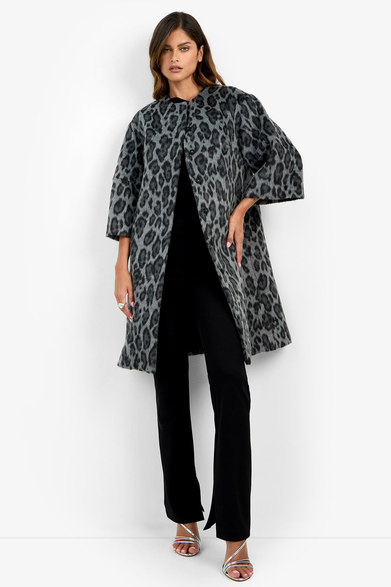 A woman models the Lovely Leopard Coat, featuring a gray and black leopard print, styled over black pants and a black top with silver heeled sandals, standing against a plain white background.