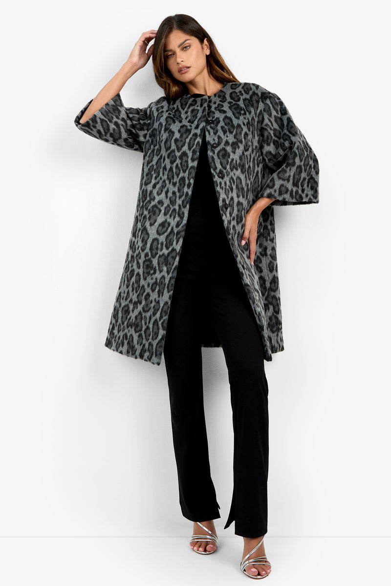 Wearing the Lovely Leopard Coat—a long, gray leopard print design—over a black outfit and silver heels, the woman stands against a plain white background with one hand touching her hair.