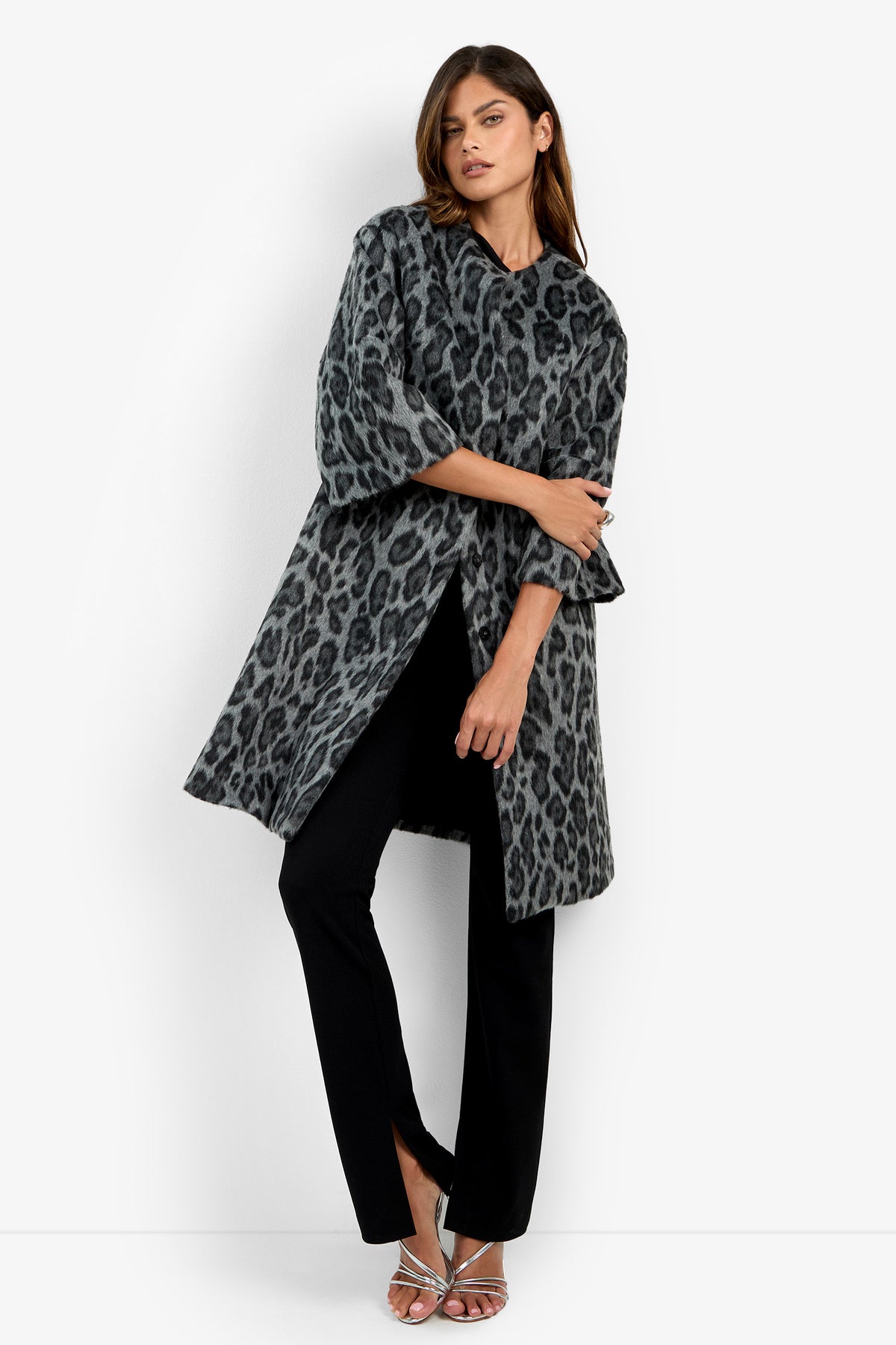 A woman models the Lovely Leopard Coat in grey leopard print, styled with black pants and silver heels. She stands against a plain white background, crossing one leg and resting an arm on her waist.