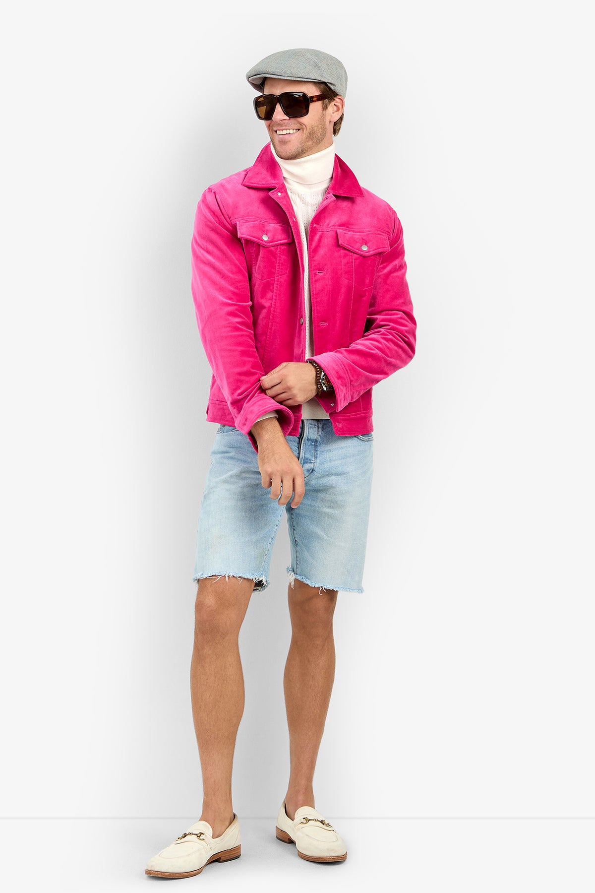 A man wearing a bright pink Angelino Trucker Jacket, white turtleneck, light-wash denim shorts, a grey cap, and sunglasses.