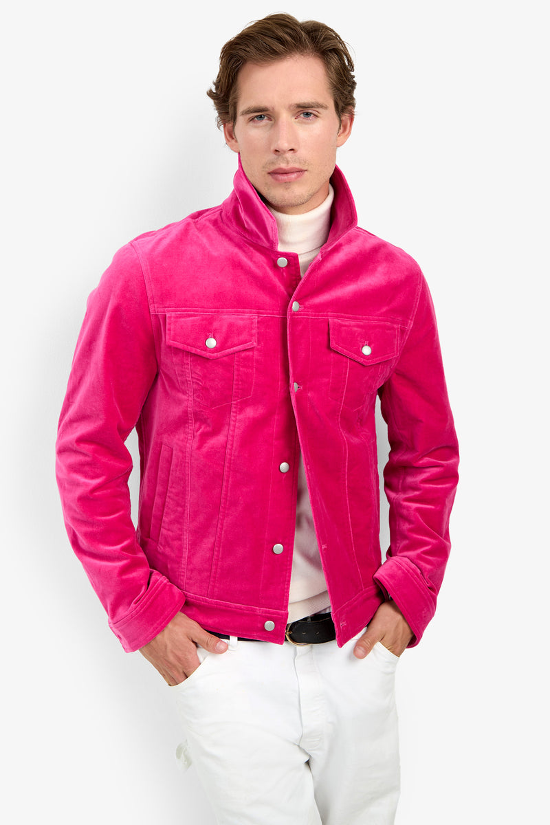 A man stands with his hands in his pockets against a plain white background, wearing the Angelino Trucker Jacket in bright pink velvet over a white turtleneck and white pants.
