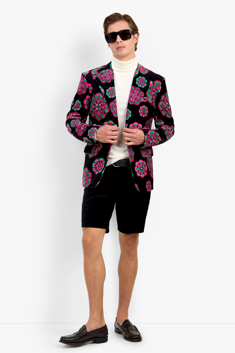 A man wearing sunglasses, a white turtleneck, black shorts, dark loafers, and the Doheny Blazer in Sautoir—a black jacket with pink and green floral patterns—stands against a plain white background.