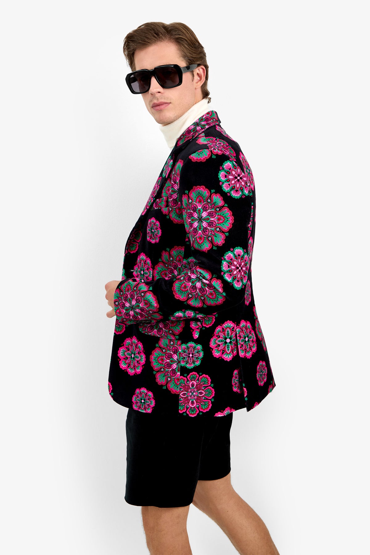 A man wearing sunglasses, the Doheny Blazer—a black suit jacket with colorful floral patterns—a white shirt, and black shorts stands against a plain white background, looking over his shoulder.