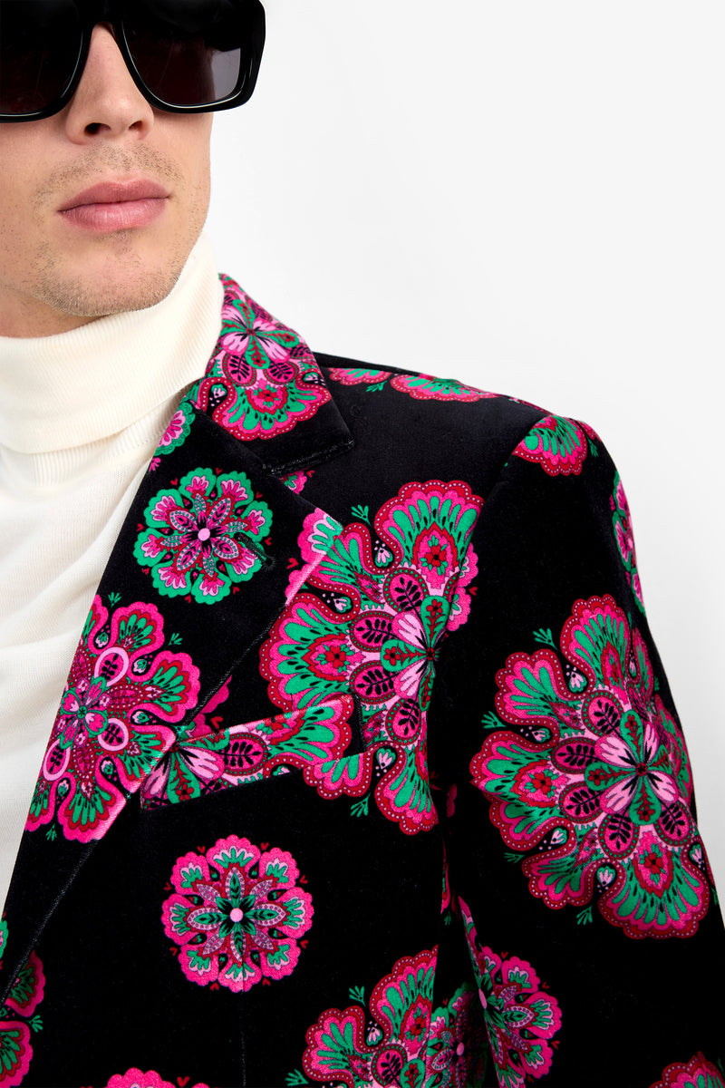 A person wearing oversized black sunglasses, a white turtleneck, and the Doheny Blazer—a velvet jacket featuring a bold pink and green floral pattern on a black background.