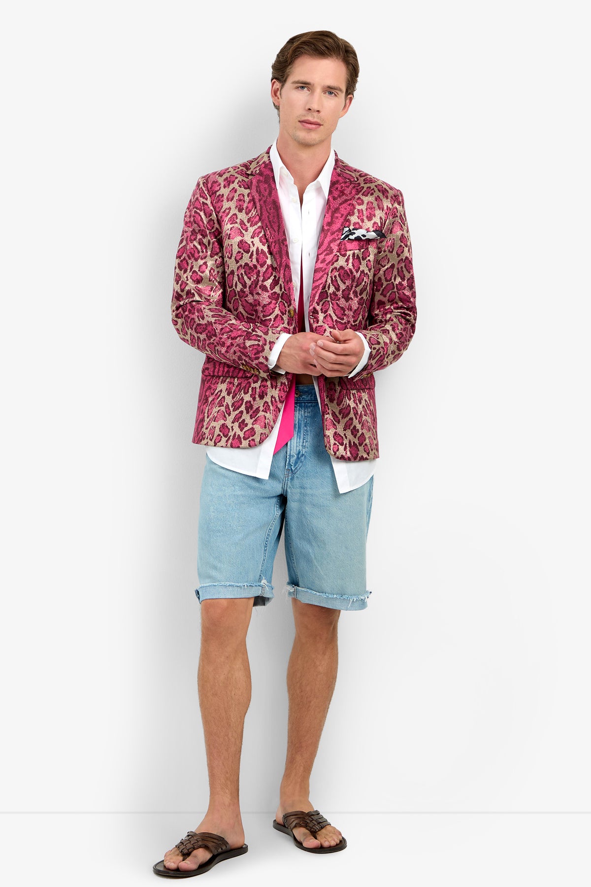 A man wearing the Thurston Blazer—a pink leopard print statement piece—paired with a white shirt, denim shorts, and sandals stands against a white background, looking at the camera with his hands clasped.