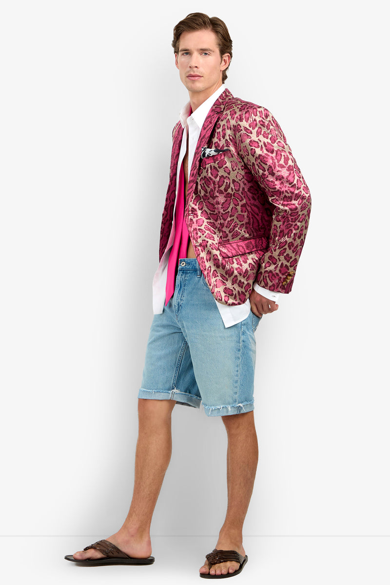 A man wearing the Thurston Blazer—featuring a bold pink leopard print—paired with a white shirt, pink tie, denim shorts, and black sandals stands against a plain white background, looking confidently at the camera.