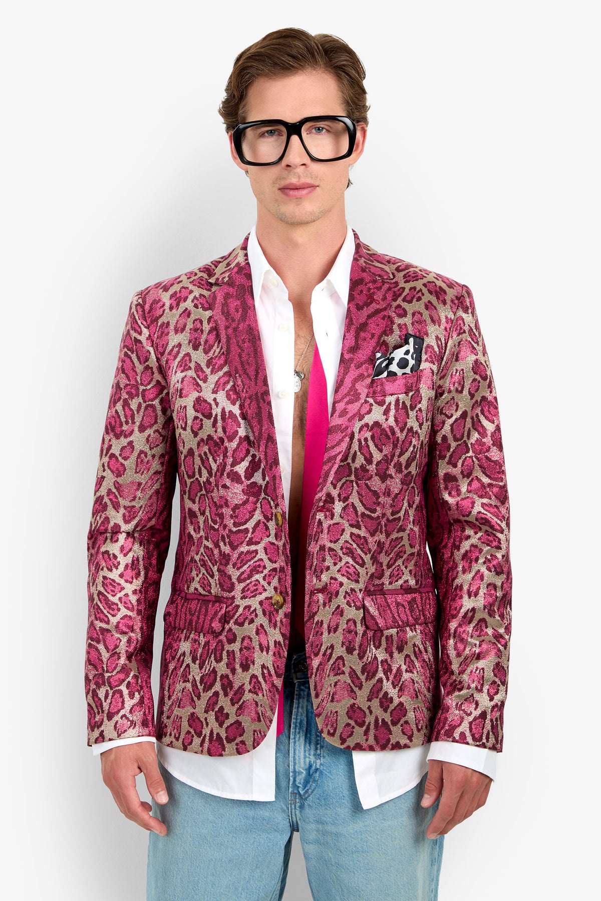 A person wearing oversized black glasses and the Thurston Blazer—a pink leopard print piece—styled over an open white shirt with a visible pink tie and blue jeans, standing against a plain white background.